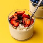 Glass jar filled with high-protein overnight oats topped with sliced strawberries and dark chocolate chunks, set against a bright yellow background